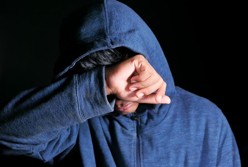 Person in blue hoodie covering face with hand, emotional distress.